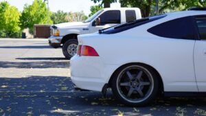 Two white vehicles parked outdoors on a sunny day - a pickup truck and a car in a parking lot with fallen leaves.