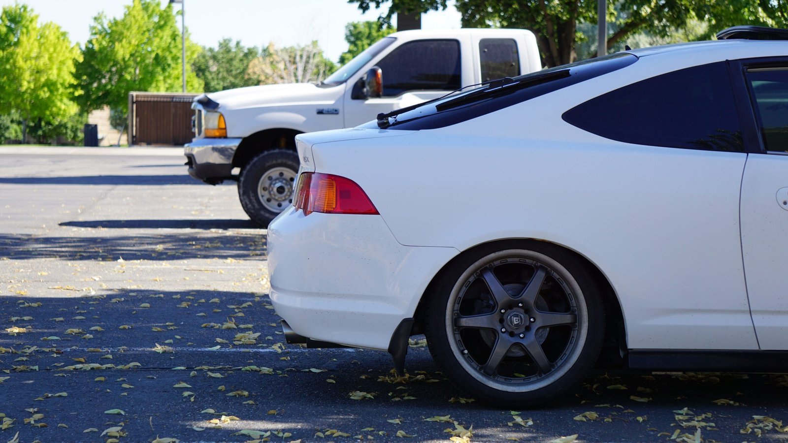 Two white vehicles parked outdoors on a sunny day - a pickup truck and a car in a parking lot with fallen leaves.